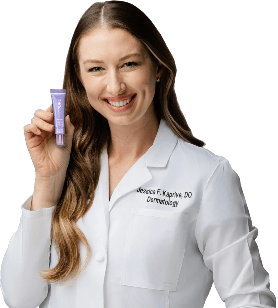 A smiling woman in a white lab coat holds a purple skincare product towards the camera. She has long, wavy brown hair and is standing against a transparent background. The lab coat has the name "Jessica F. Kaprive, DO" and the word "Dermatology" embroidered on it. Her expression is friendly and approachable, suggesting expertise in skincare.