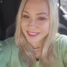 Smiling woman with long blonde hair sitting in a car, wearing a light green sweater and a heart-shaped necklace with blue stones.
