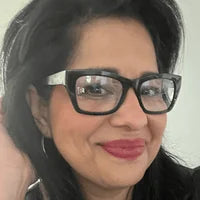 A woman with long dark hair and wearing large black glasses smiles at the camera. She is lightly touching her hair with one hand and has a warm, friendly expression. The background is softly lit, creating a bright and inviting atmosphere.