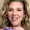 A smiling person with long, wavy blonde hair holds a small purple container up to the camera. They appear cheerful and engaged, set against a plain background.